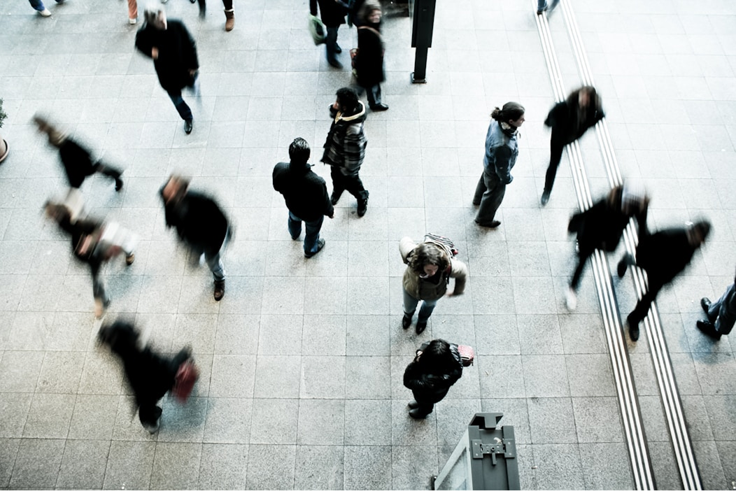 Blurred crowd moves in various directions on a busy sidewalk, conveying motion and urban hustle. A few people stand still amid the bustling scene.