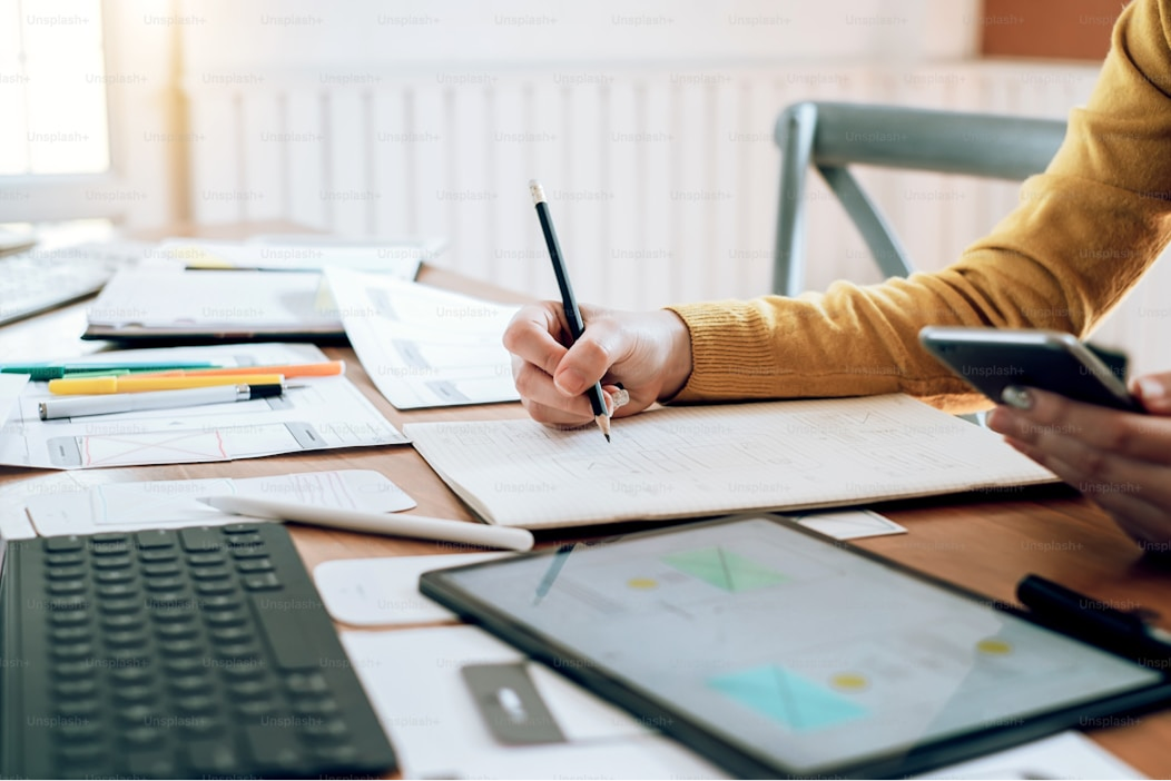 Close-up of a person in a yellow sweater writing on a notepad with a pen, surrounded by papers, a tablet, and a keyboard, conveying focus and productivity.