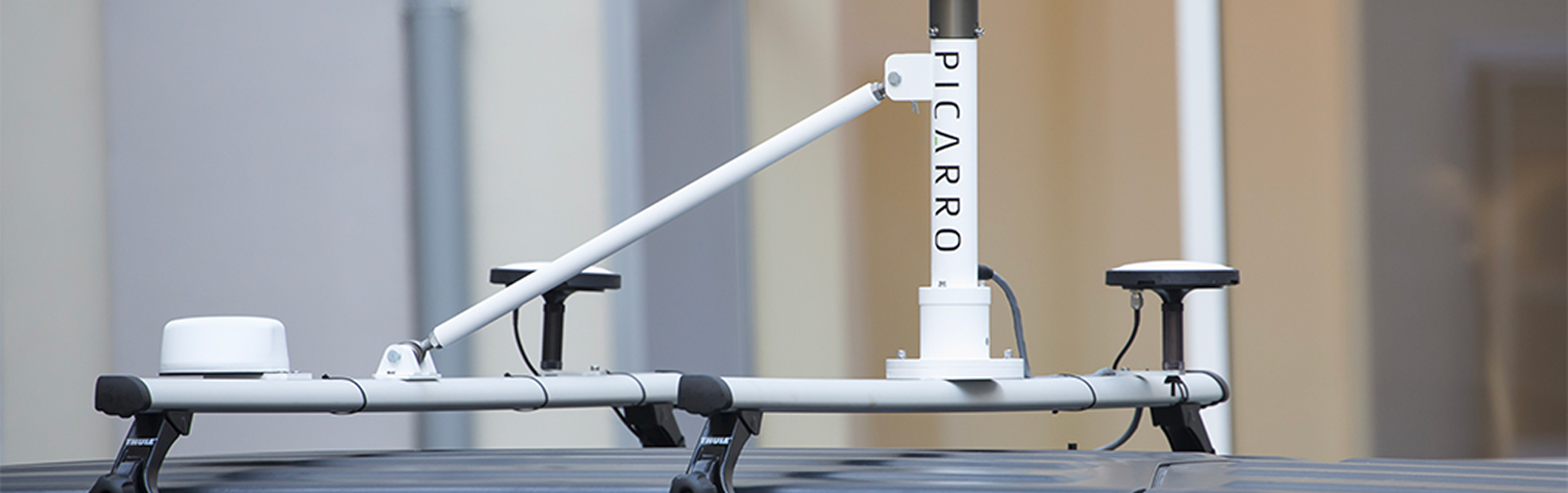 Rooftop-mounted Picarro air quality sensors with white poles and circular antennas, set against a blurred urban backdrop. High-tech and precise.