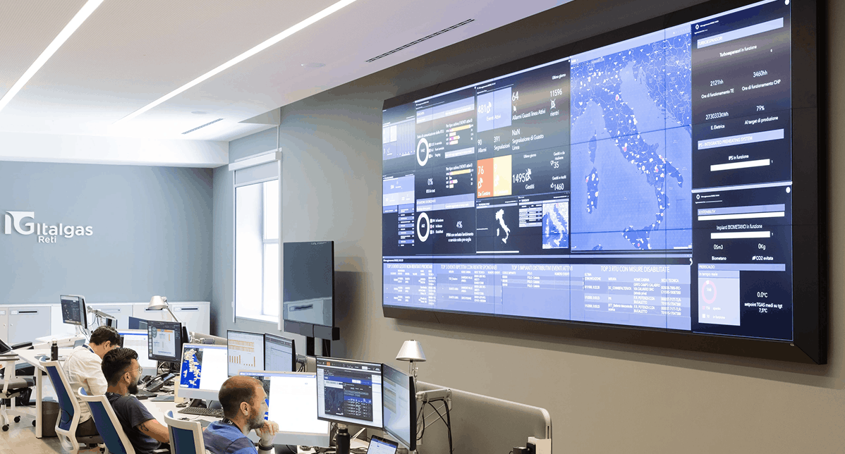 Modern control room with large digital displays showing data and maps. Several people work at desks with computers, creating a focused atmosphere.