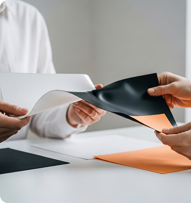 Two people examining samples, holding black and white sheets over a white table with orange and black papers. The scene conveys collaboration and focus.
