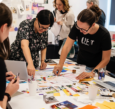 A group of people collaborates energetically at a table covered with colorful papers, photos, and laptops, conveying creativity and teamwork in a lively workspace.