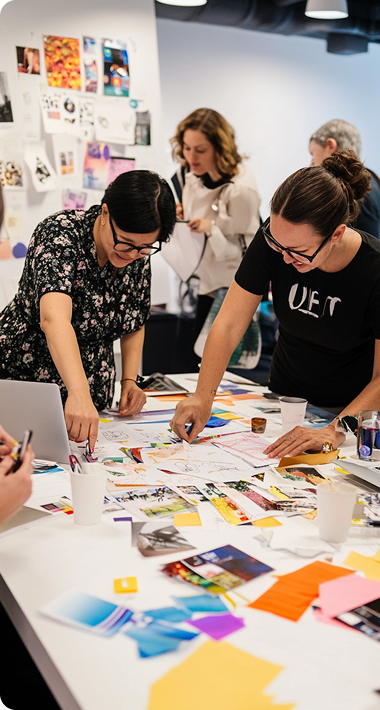 A group of people collaborates energetically at a table covered with colorful papers, photos, and laptops, conveying creativity and teamwork in a lively workspace.