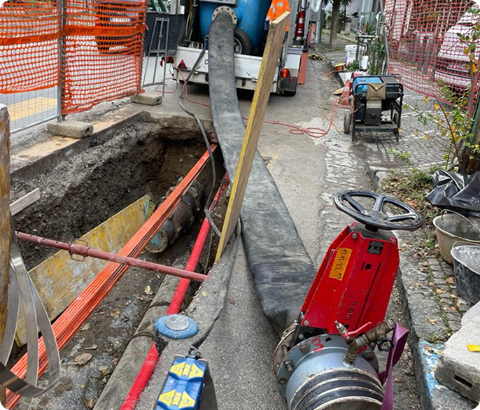 Construction site with a large trench on the left, surrounded by orange safety barriers. Various construction equipment and cables are present.