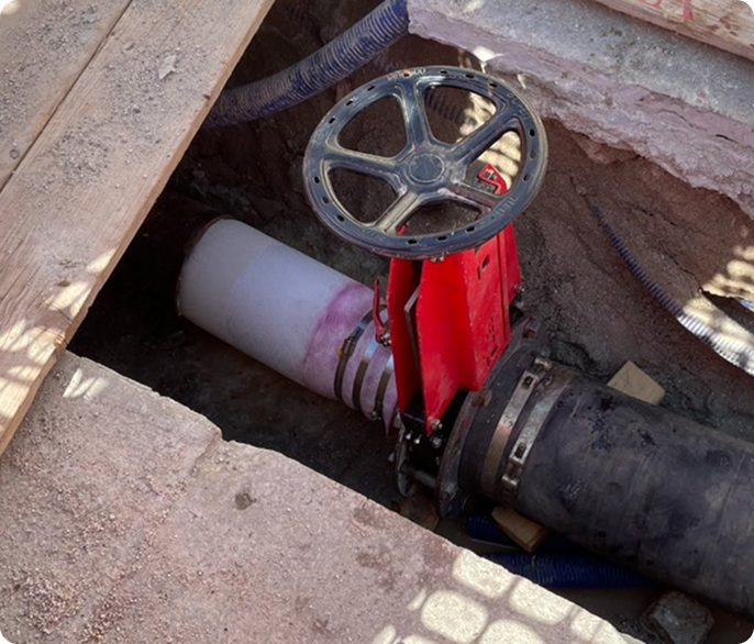 A construction site close-up showing a red valve with a large wheel control, connected to black and white pipes. The setting is rugged and industrial.
