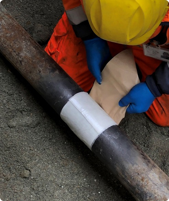 Worker in a yellow helmet and orange overalls repairs an underground pipe. They wear blue gloves and apply a patch. The scene suggests construction work.