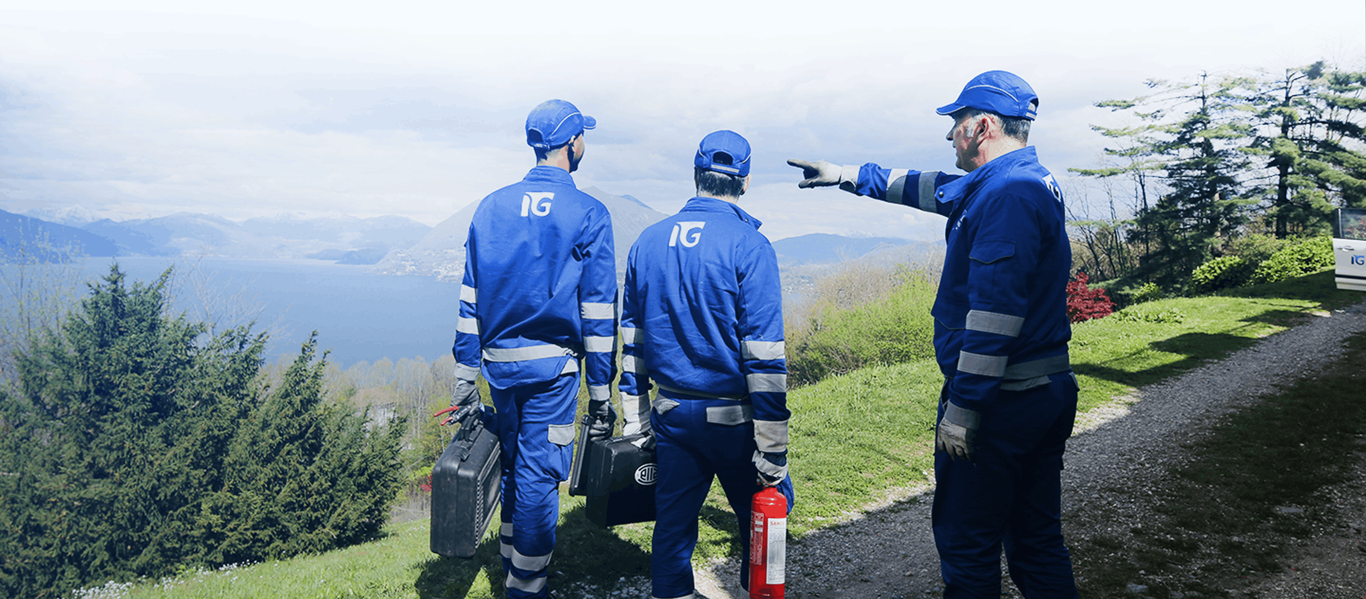 Three workers in blue uniforms and caps stand on a scenic hillside overlooking a lake, with one pointing toward the mountains. They carry safety equipment, creating a sense of teamwork and focus.